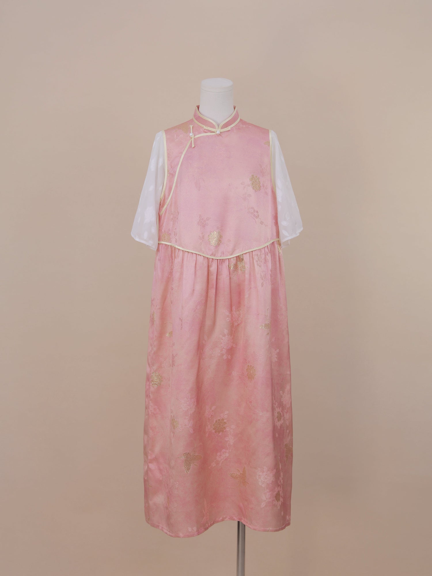 Full front view of a pink silk handmade qipao with gold floral jacquard, mandarin collar, pearl frog buttons, and mid-calf length skirt with sheer white sleeves.