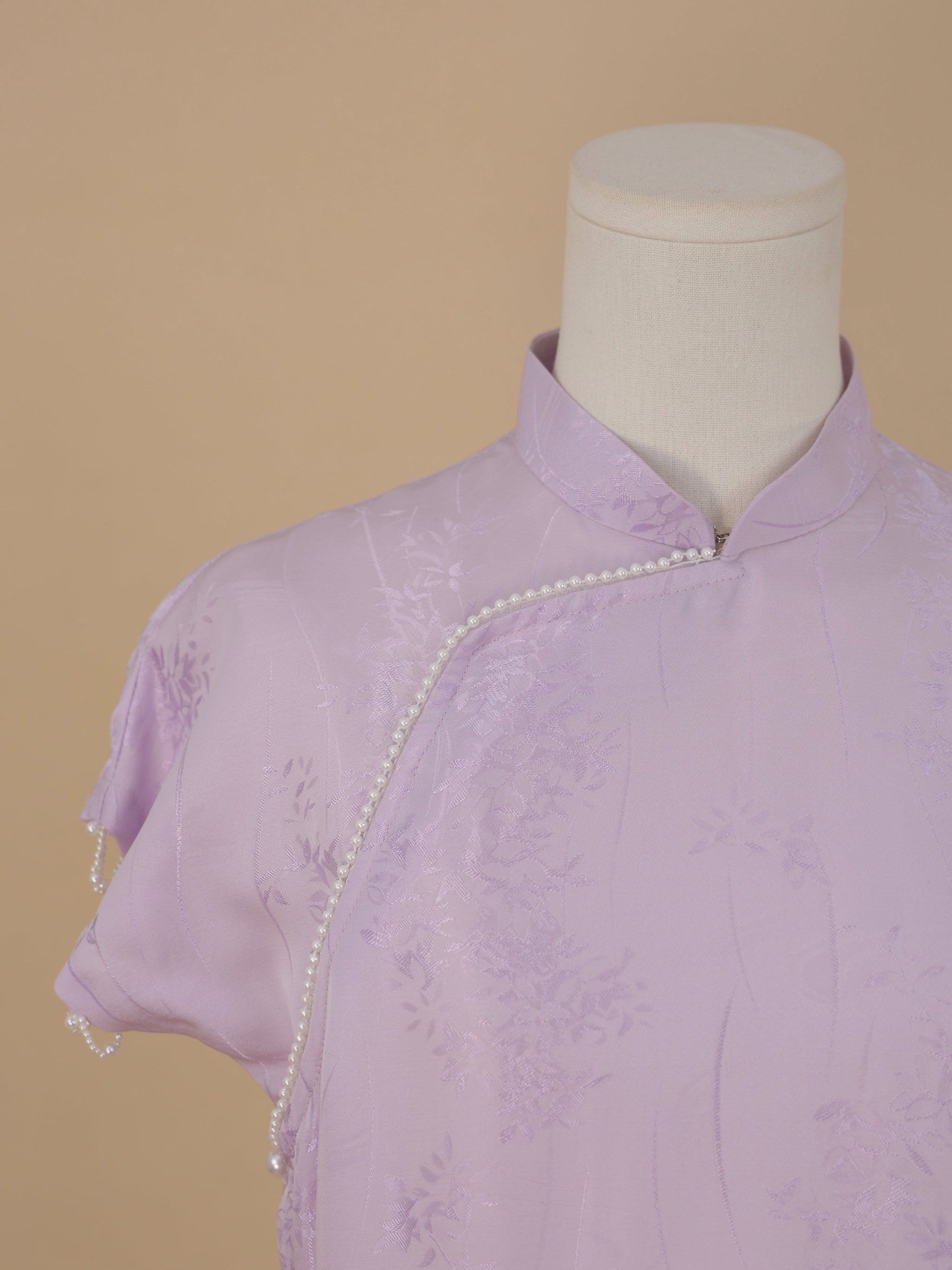 Detailed view of the mandarin collar and pearl-embellished edge of a lavender silk qipao with subtle floral patterns.