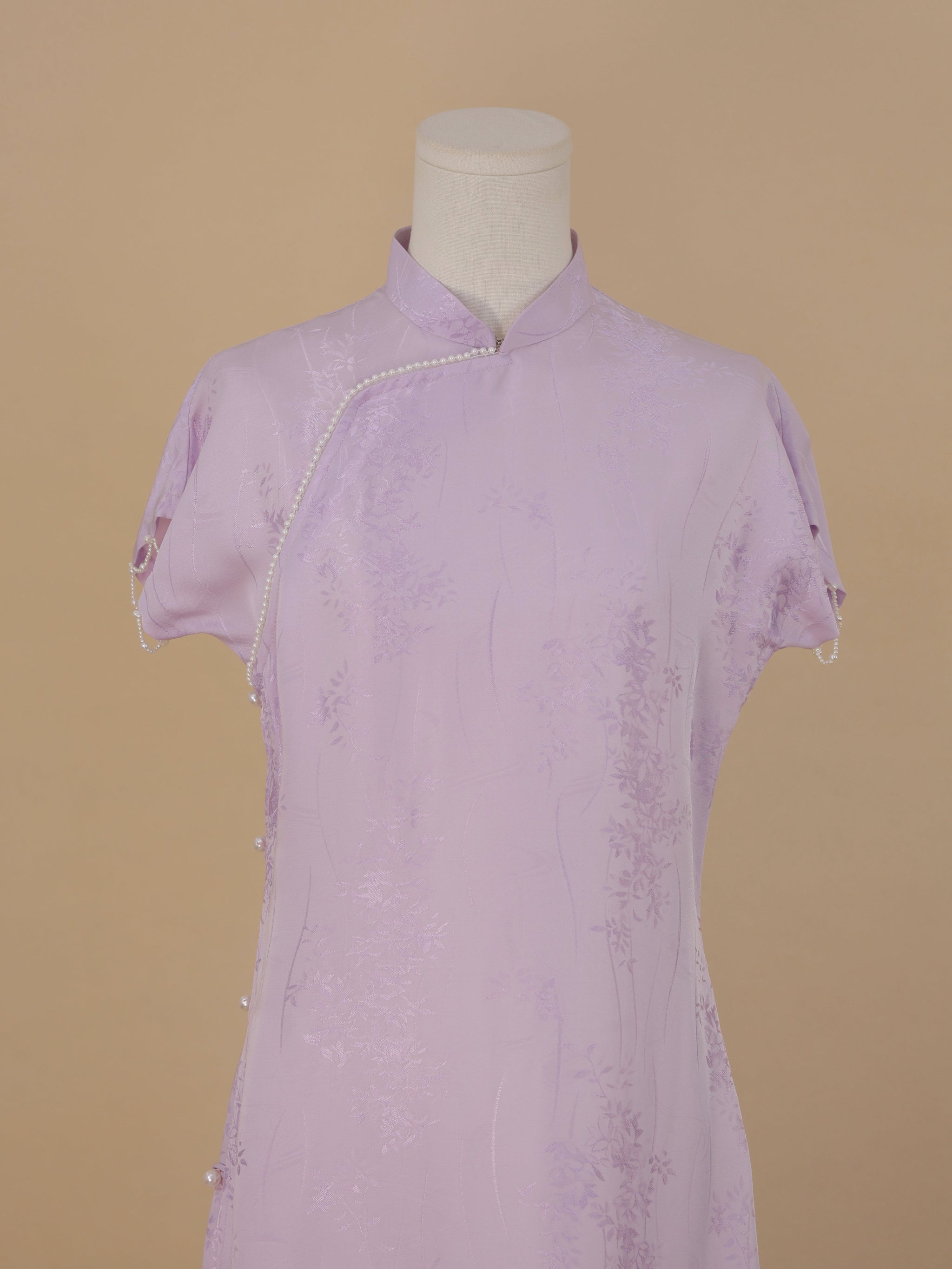 Close-up view of a lavender silk qipao featuring delicate floral jacquard patterns and pearl trim along the asymmetrical front closure.