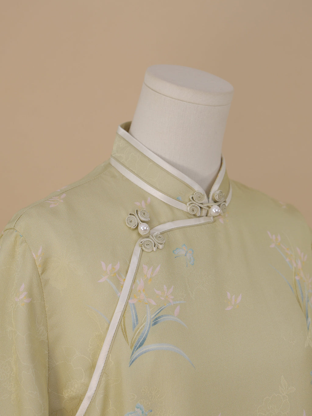 Close-up of the diagonal front detail on a pale sage green qipao, with white piping, flower-shaped frog buttons with pearl accents, and fabric featuring blue butterflies and pink flowers.