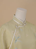 Close-up of the diagonal front detail on a pale sage green qipao, with white piping, flower-shaped frog buttons with pearl accents, and fabric featuring blue butterflies and pink flowers.