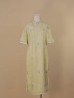 Full front view of a pale sage green short-sleeve qipao with a straight silhouette, diagonal white piping design, and fabric adorned with pink flowers and blue butterflies.