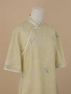 Close-up of a pale sage green silk short-sleeve qipao with soft pink floral and blue butterfly motifs, featuring white piping along the diagonal front and pearl-embellished frog buttons.