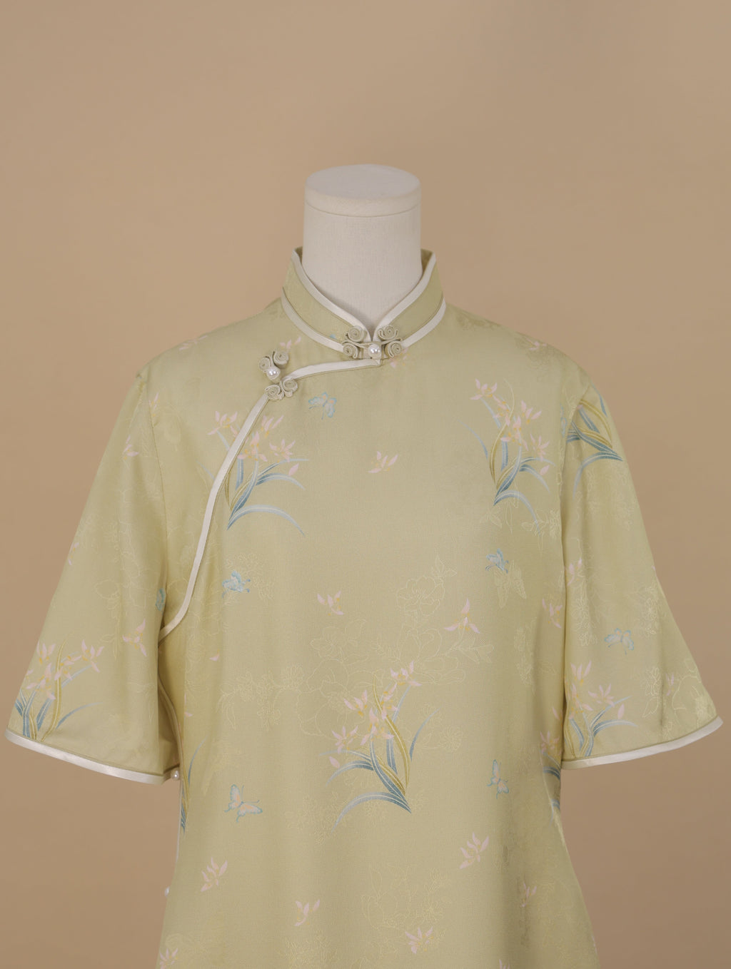 Upper front view of a pale sage green short-sleeve qipao with a stand collar, white trim, and pearl frog buttons, decorated with soft pink flowers and blue butterfly motifs.