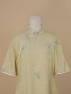 Upper front view of a pale sage green short-sleeve qipao with a stand collar, white trim, and pearl frog buttons, decorated with soft pink flowers and blue butterfly motifs.