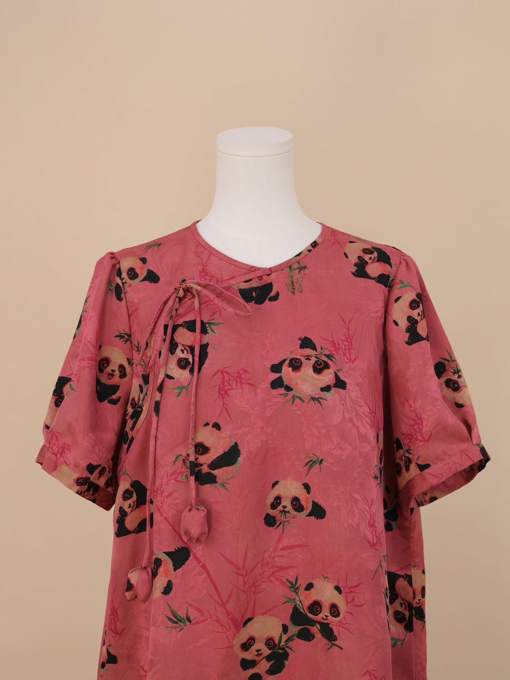 Front half-body view of a pink panda print short-sleeve dress, with a draping tie at the neckline and a relaxed sleeve design, fully covered in panda and bamboo prints.