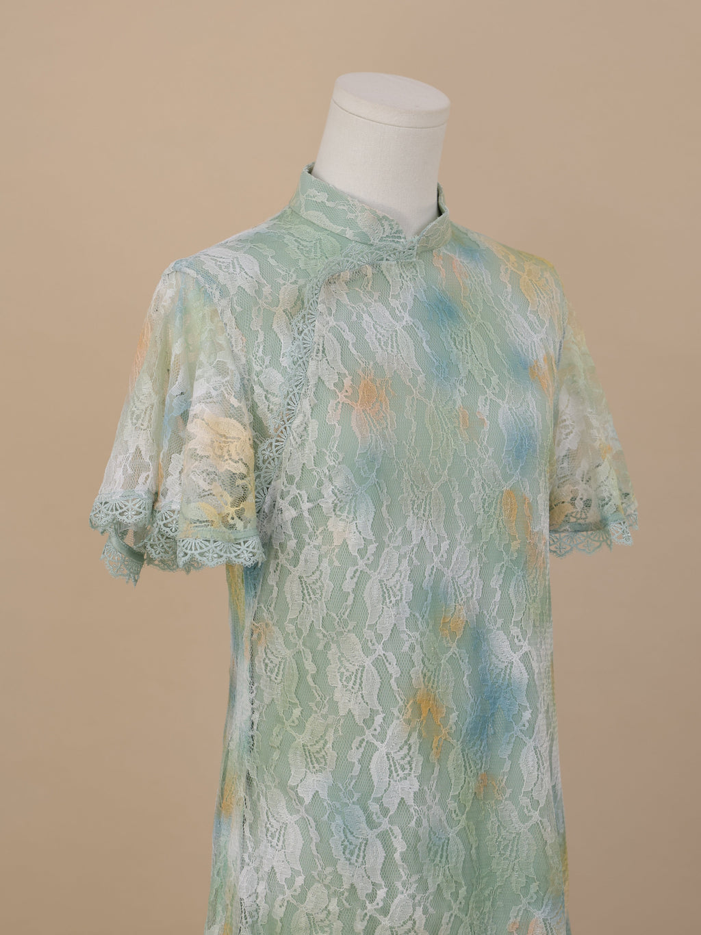 Angled close-up of the upper body of a sea green lace cheongsam, featuring short sleeves, a stand collar, and white floral lace with yellow and blue gradient patterns.