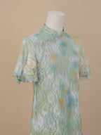 Angled close-up of the upper body of a sea green lace cheongsam, featuring short sleeves, a stand collar, and white floral lace with yellow and blue gradient patterns.