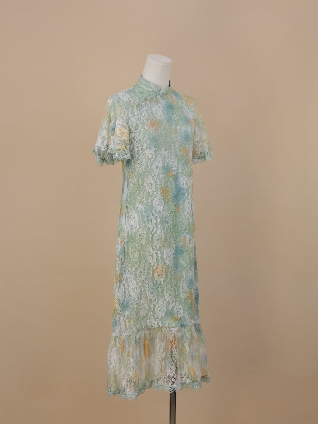 An angled full-body view of a sea green lace cheongsam with yellow and blue gradient floral lace, short sleeves, a stand collar, and a fishtail ruffle hem.