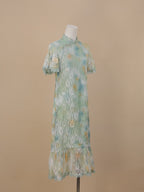 An angled full-body view of a sea green lace cheongsam with yellow and blue gradient floral lace, short sleeves, a stand collar, and a fishtail ruffle hem.