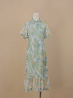 A sea green lace cheongsam with yellow and blue gradient floral patterns, short sleeves, stand collar, and fishtail ruffle hem, displayed against a beige background.