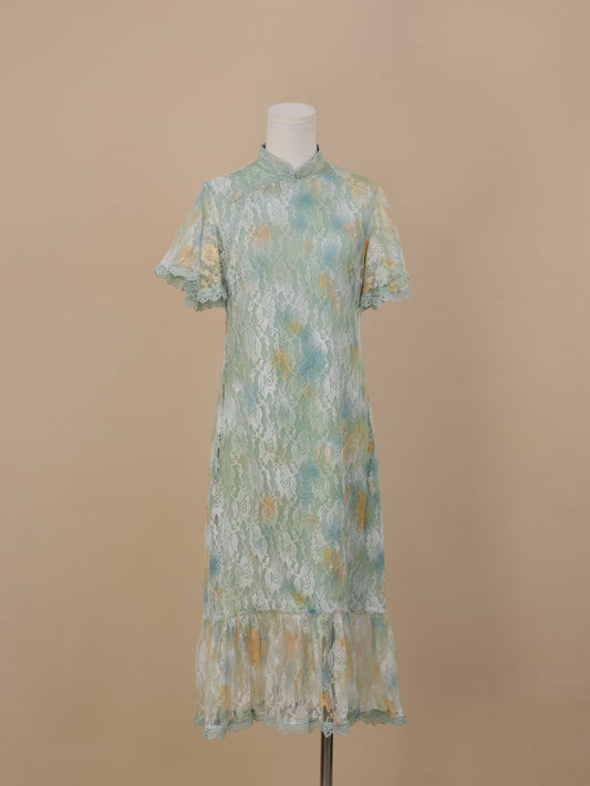 A sea green lace cheongsam with yellow and blue gradient floral patterns, short sleeves, stand collar, and fishtail ruffle hem, displayed against a beige background.