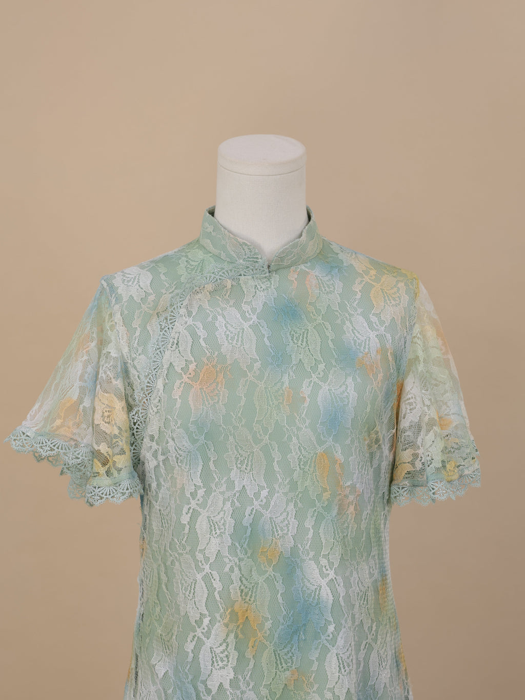 Close-up of the upper front of a sea green lace cheongsam with yellow and blue gradient floral accents, featuring short sleeves and a stand collar, set on a beige background.