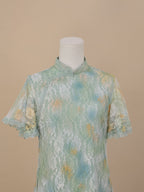 Close-up of the upper front of a sea green lace cheongsam with yellow and blue gradient floral accents, featuring short sleeves and a stand collar, set on a beige background.