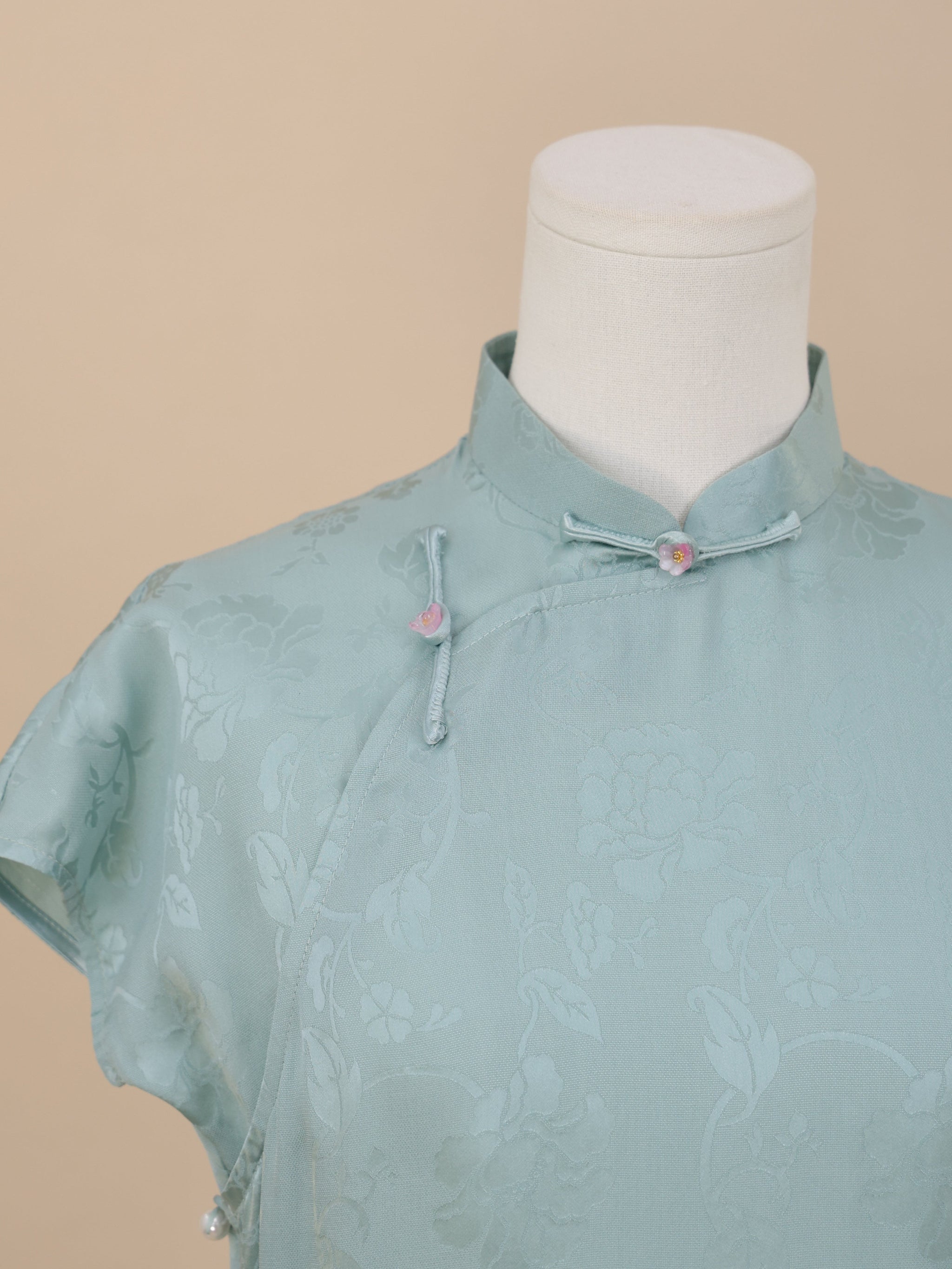 Close-up of the neckline of a Celadon Blue silk cheongsam highlighting the floral jacquard fabric, Mandarin collar, and pink flower knot buttons.