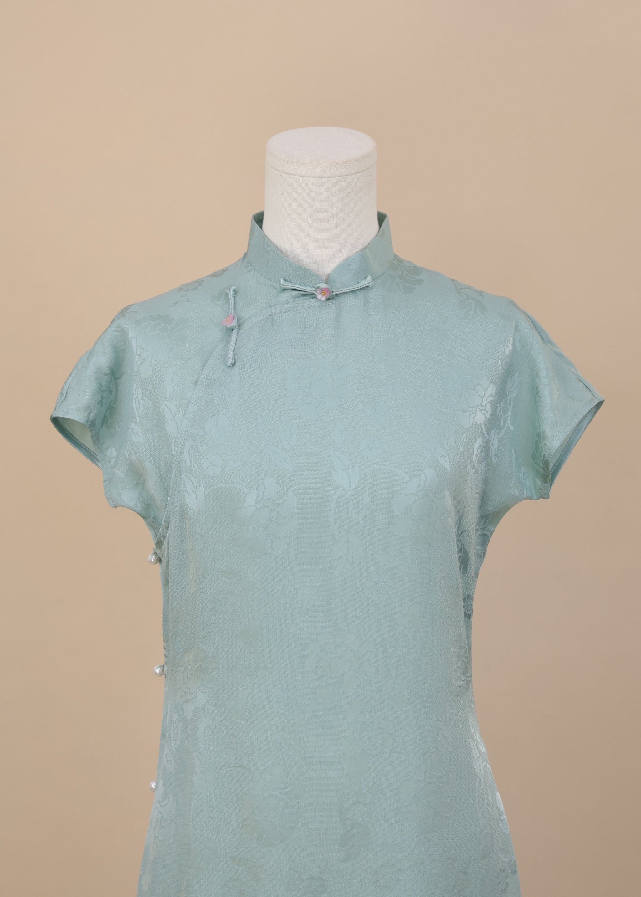 Frontal view of the upper body of a Celadon Blue silk cheongsam with short sleeves, Mandarin collar, floral jacquard weave, and decorative pink knot buttons.